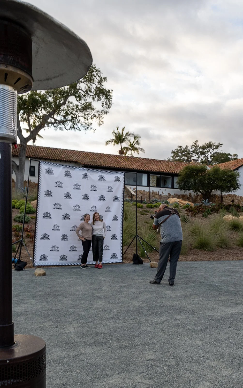 Two women posing for a photo in front of a backdrop with logo and text, with a photographer taking their picture, set outdoors with desert plants and a building in the background.