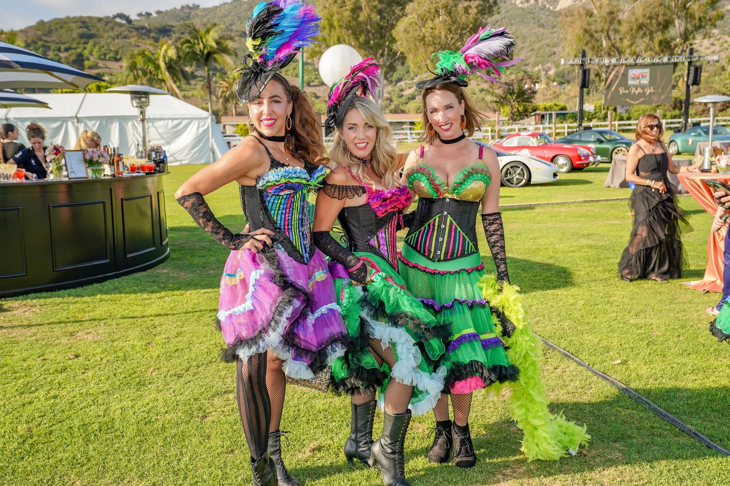 Three women dressed in colorful carnival costumes with feathers and lace, posing outdoors at a festive event.