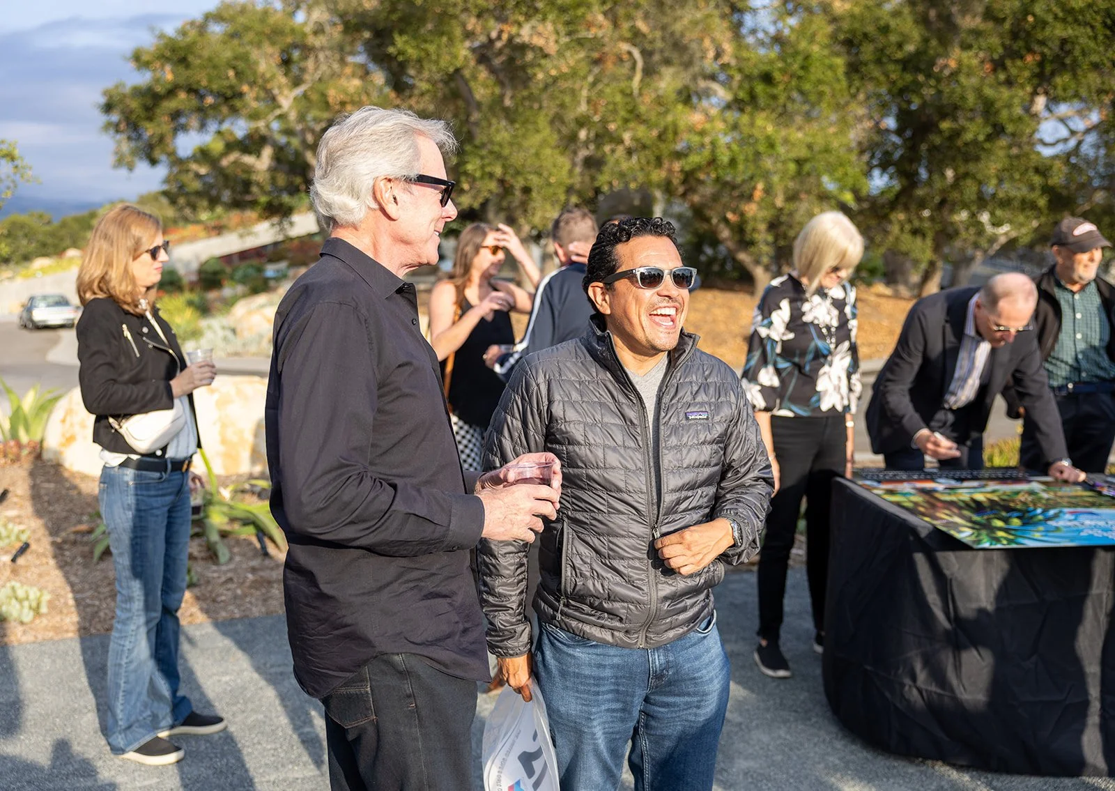 Group of people gathered outdoors, socializing, with two men in the foreground laughing and talking.
