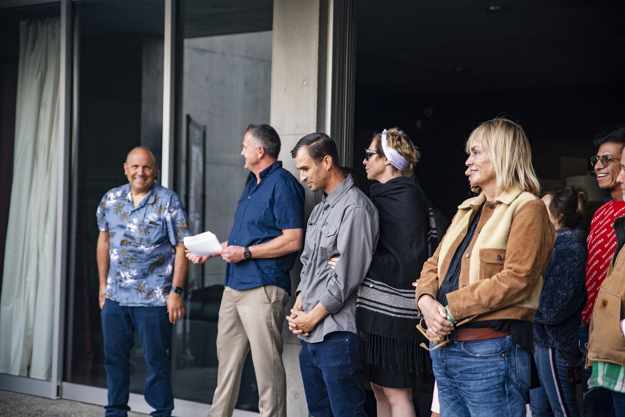 Group of people standing in a line outside a building, some smiling and engaged in conversation.