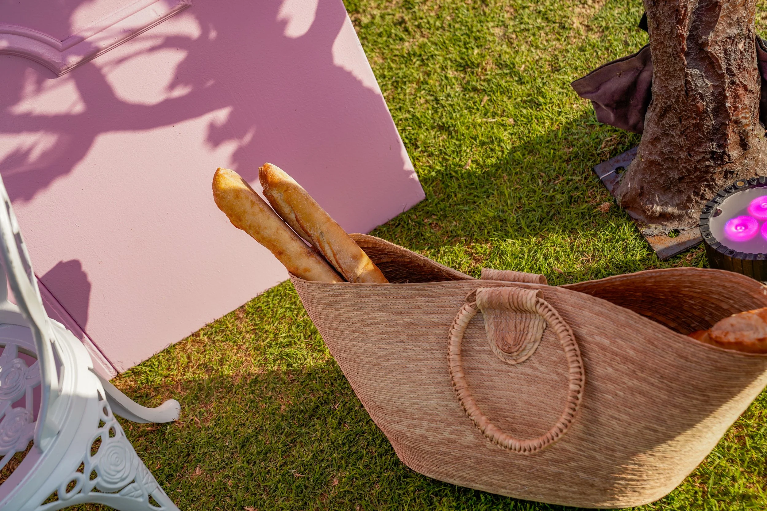 Brown woven tote bag with two breadsticks inside, placed on green grass near a pink and white decorative chair and a small round table with purple candles in a black container. A brown fabric is wrapped around the base of a tree on the right side.