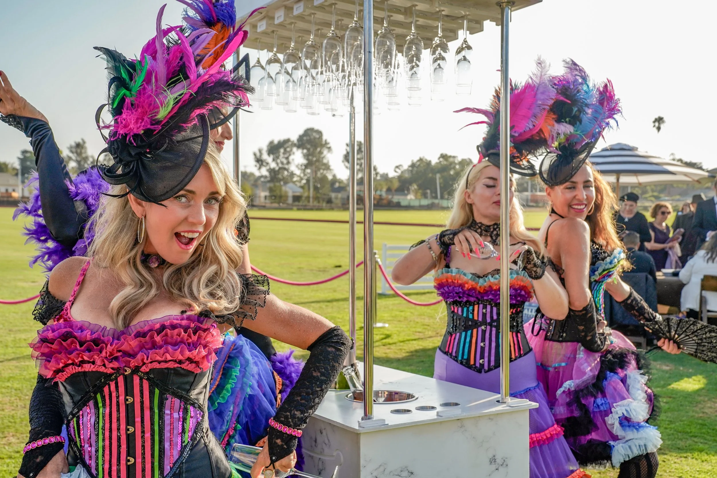 Three women in colorful, elaborate costumes with feathered hats at an outdoor event, with wine glasses hanging above a bar.