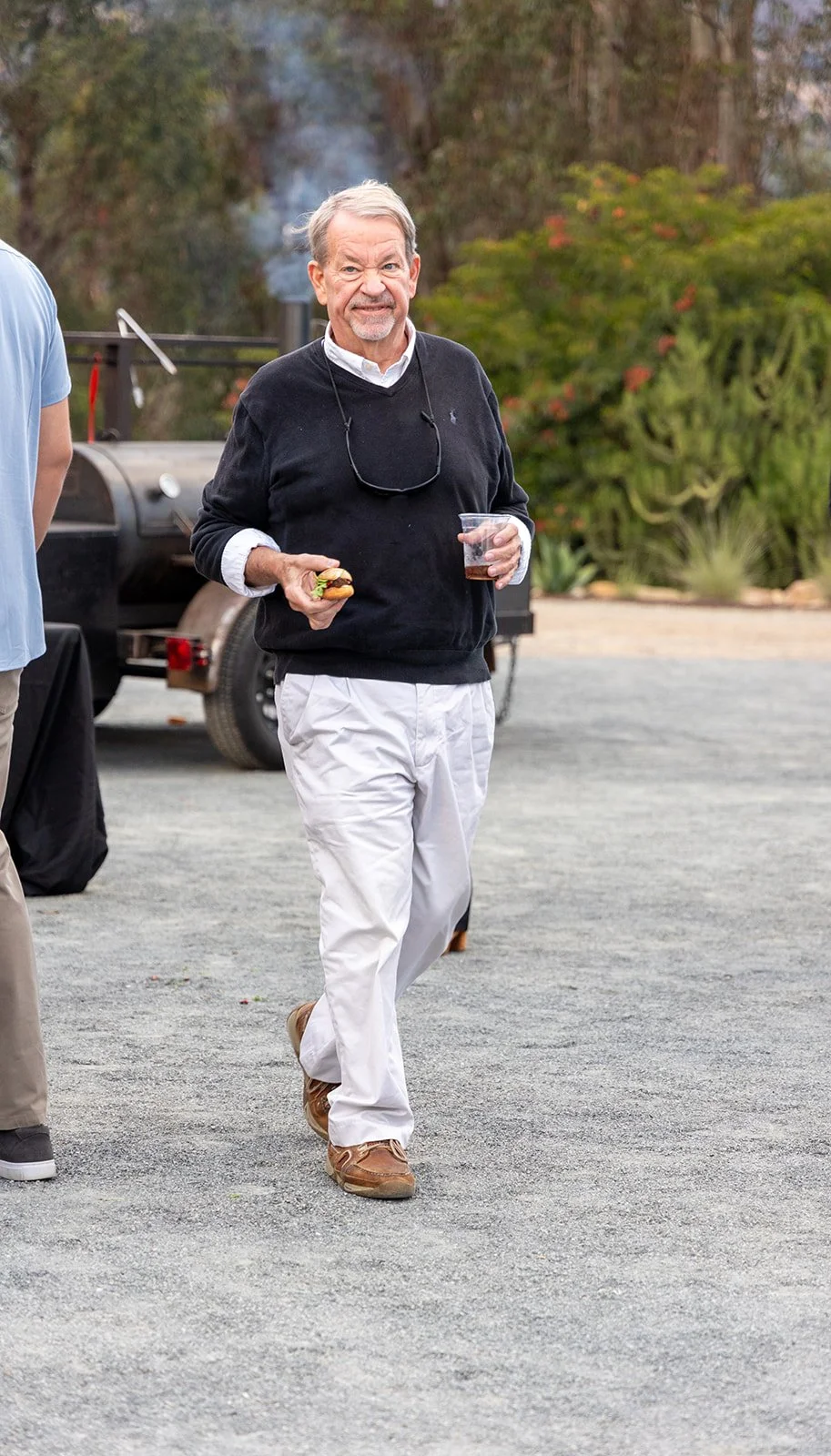 An older man with gray hair and a beard, wearing a black sweater over a white collared shirt, and white pants, holding a hot dog in one hand and a plastic cup in the other, walking outdoors near a black barbecue grill.