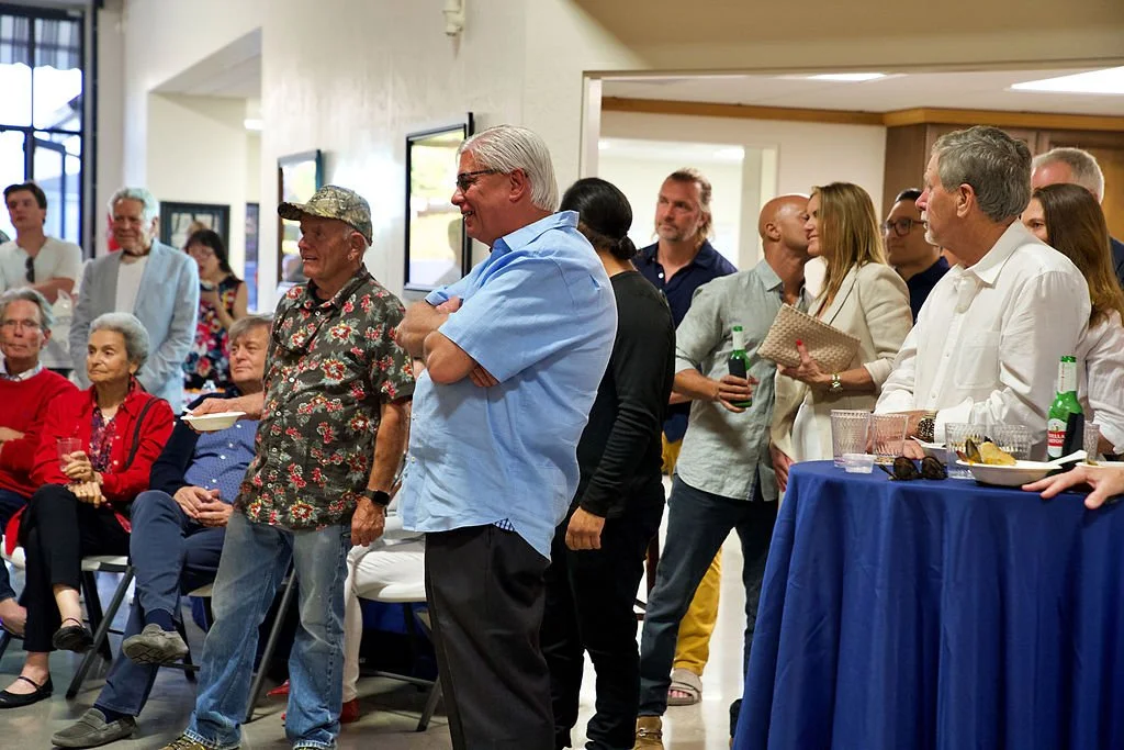 A group of people gathered at an indoor event, with some standing and some seated, engaging in conversations and enjoying refreshments.