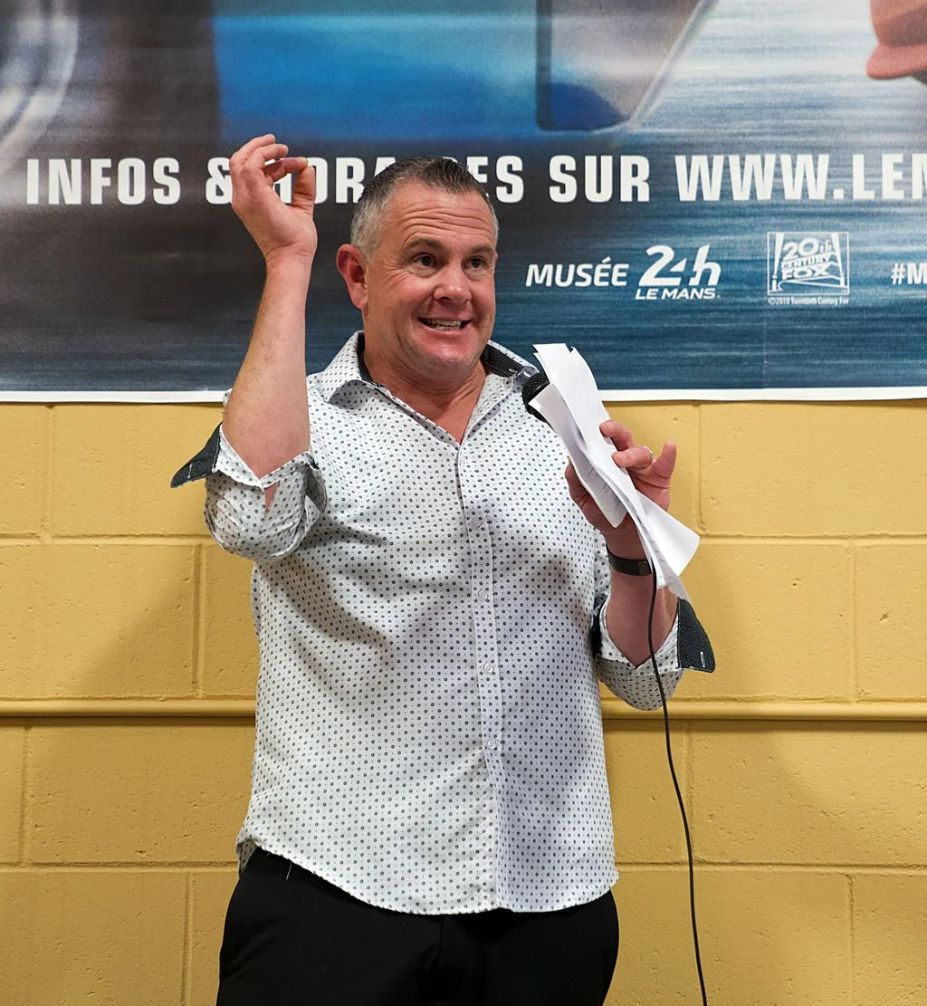 A man speaking into a microphone, holding papers, smiling, in front of a yellow wall and a large blue banner.
