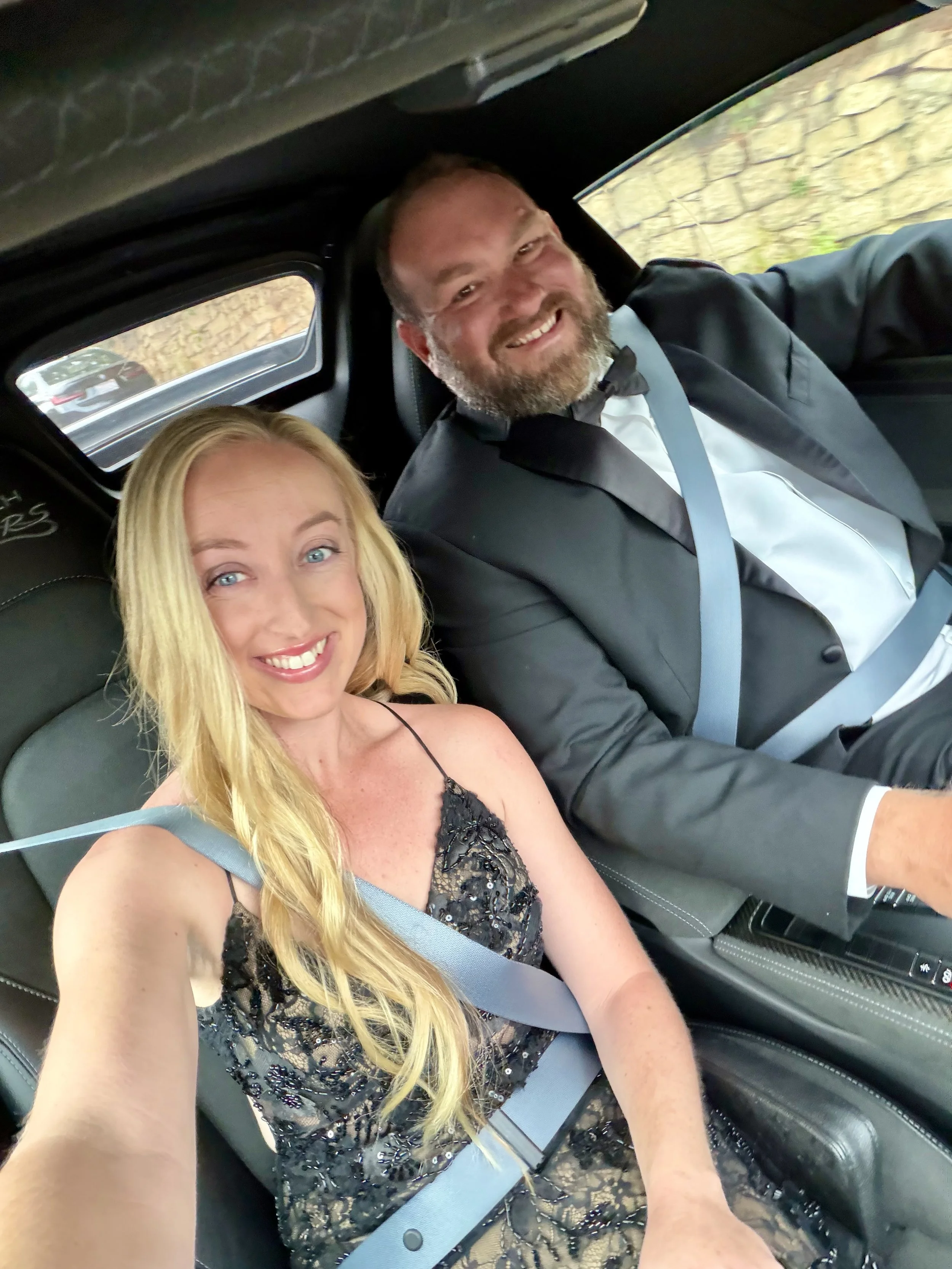 blonde-woman-in-black-dress-and-man-in-tuxedo-smiling-in-car--025e8035.jpeg