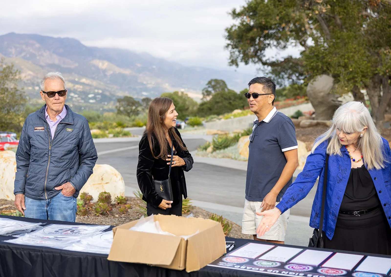 People standing outdoors in a conversation near a table with event materials, with mountains and trees in the background.