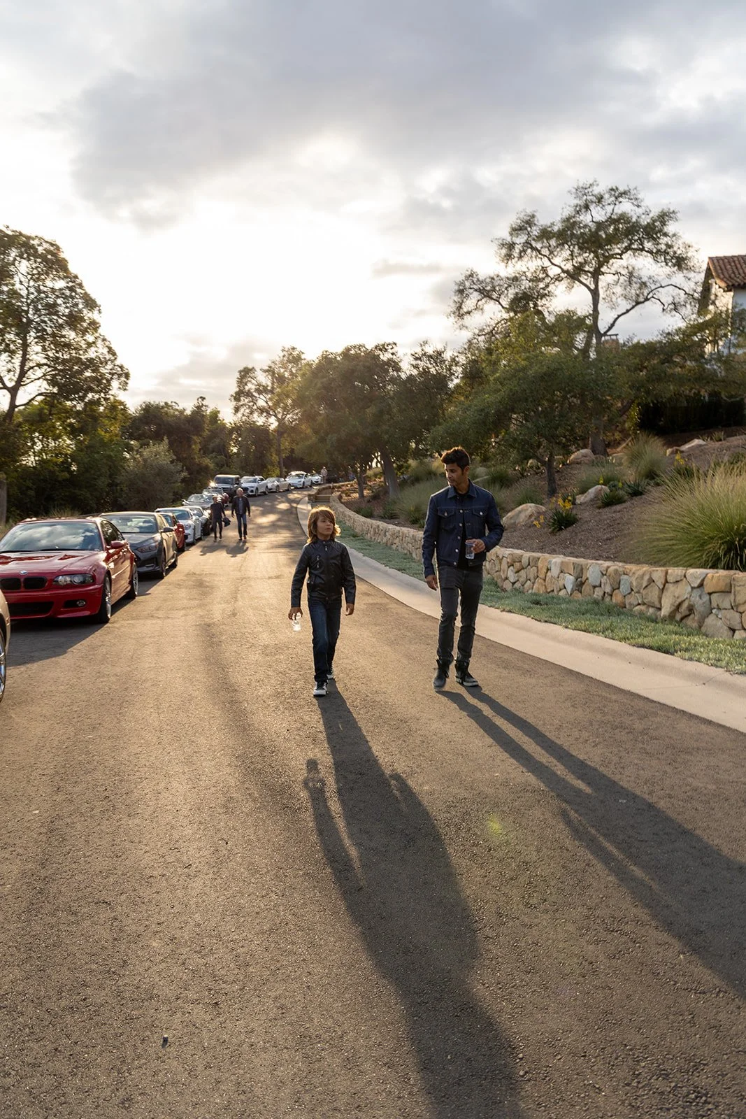 A man and a girl walking on a paved road in a residential area during sunset, with parked cars on the side and trees with landscaped bushes nearby.