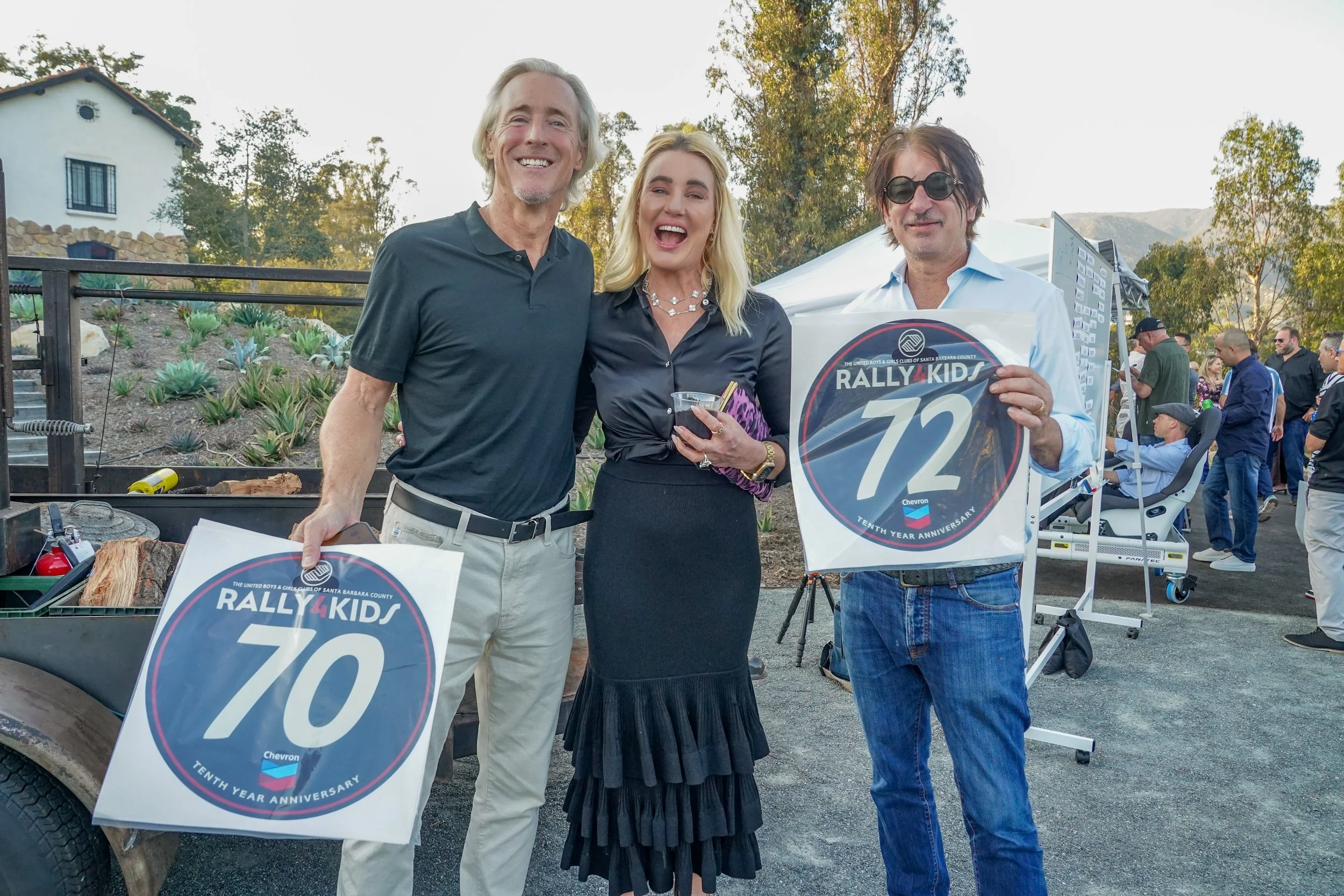 Three people smiling at an outdoor event, holding rally signs with numbers 70 and 72, and standing near a cart with tools. The background includes other people, trees, and a white tent.