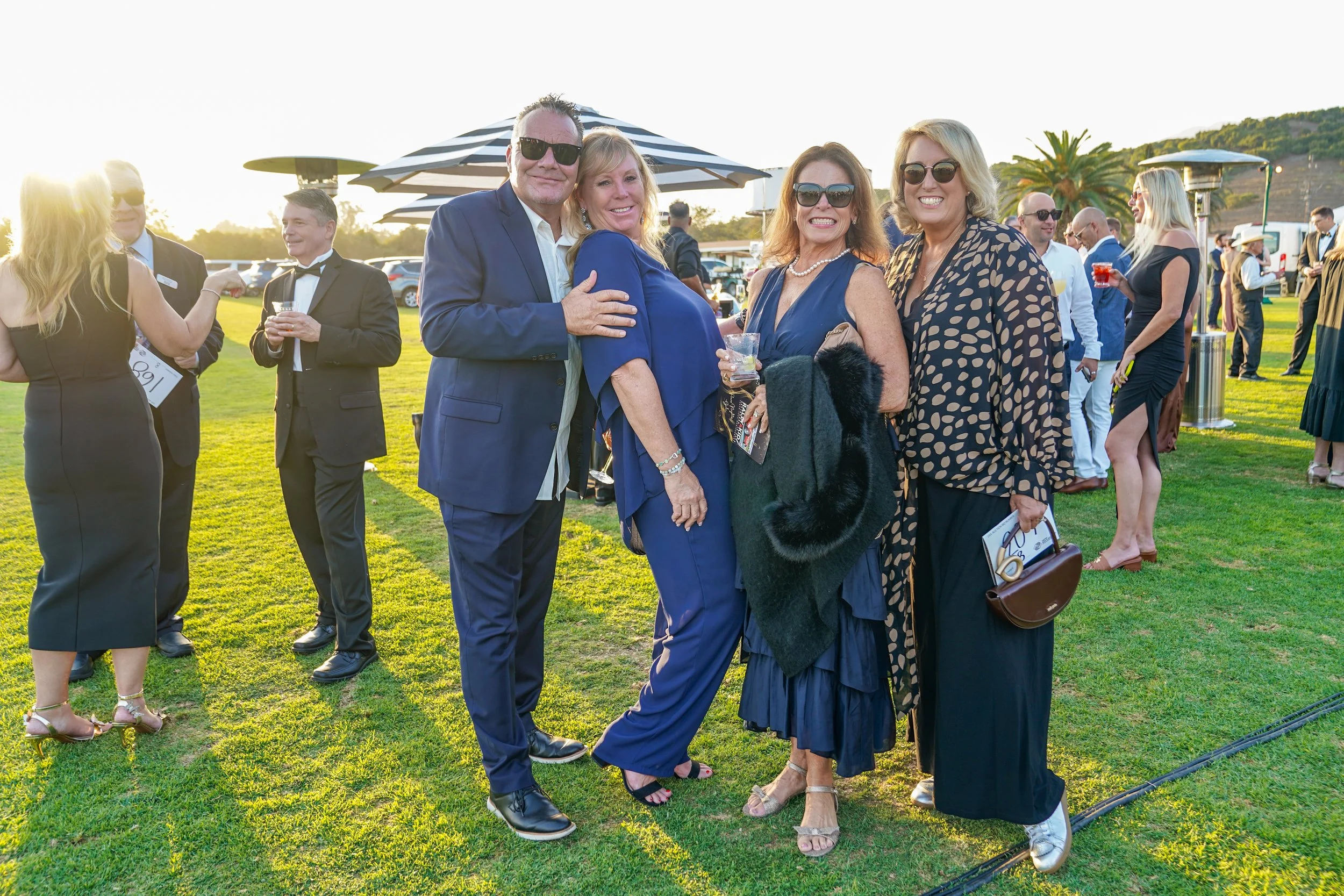 Group of people dressed in formal attire, standing outdoors at a sunny event, smiling and holding drinks.