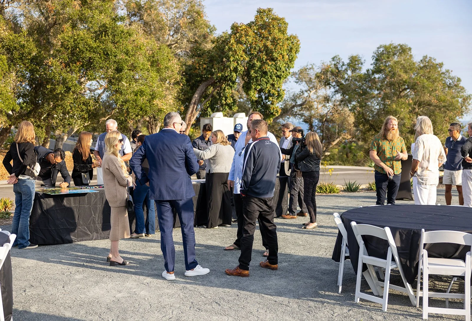 Group of people gathered outdoors around tables and chairs, socializing in a setting with trees and a road in the background, during the daytime.
