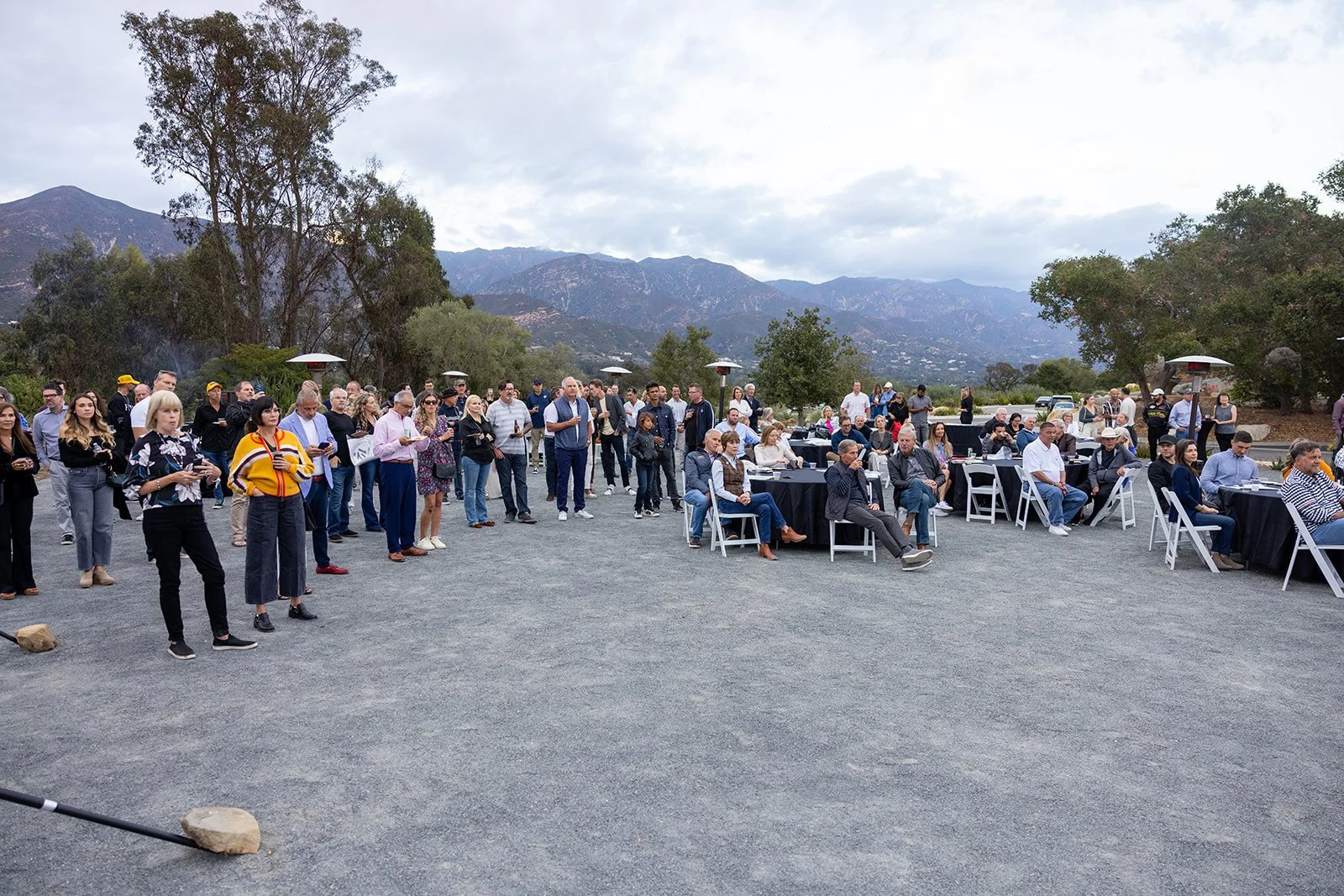 A large group of people gathered outdoors at an event, with some standing and others seated at black-covered tables with white chairs, in a mountainous area with trees and cloudy sky in the background.