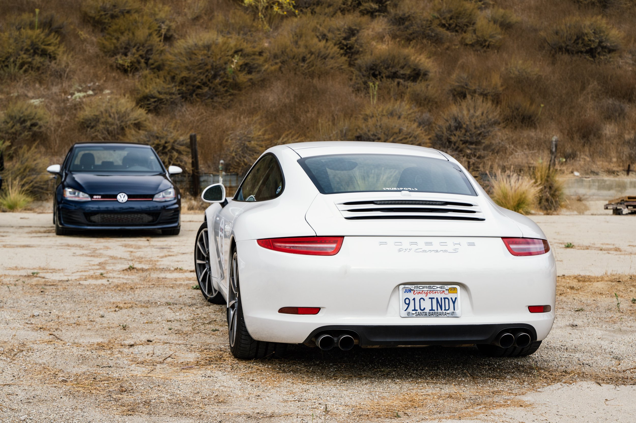 A white Porsche 911 Carrera S with a California license plate reading 91C INDY, parked in a dirt lot with a black Volkswagen Golf R in the background and dry, brown shrubbery on a hillside.