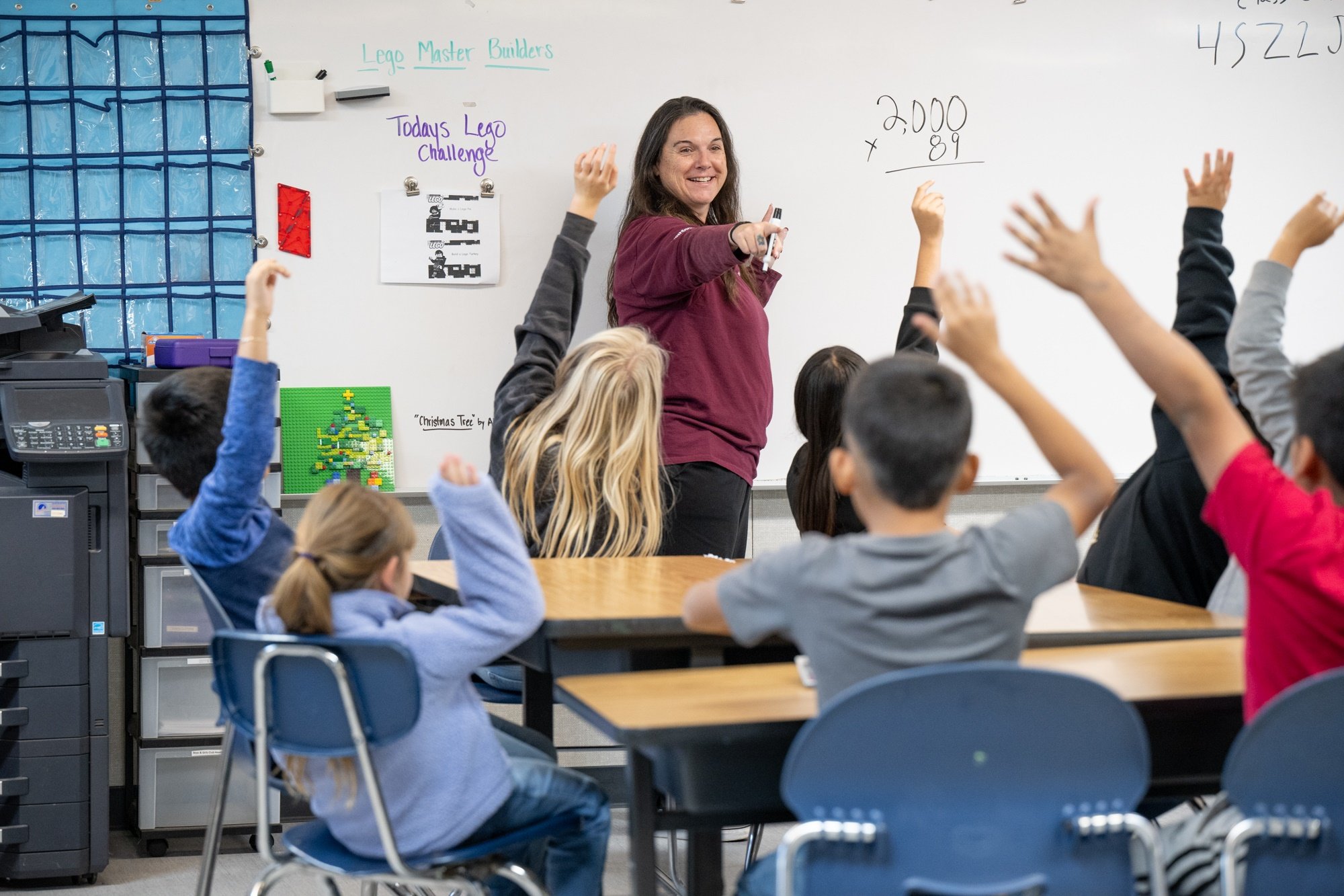 A classroom scene with students raising their hands, and a teacher standing at the whiteboard smiling. The whiteboard has writing and math problems on it, and there is a colorful Lego Christmas tree on the side.