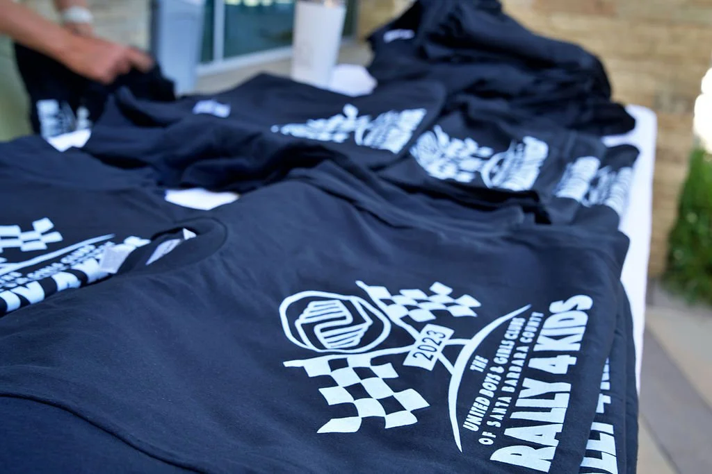 Tables with black T-shirts featuring a racing event logo, laid out outdoors with a brick wall and greenery in the background.