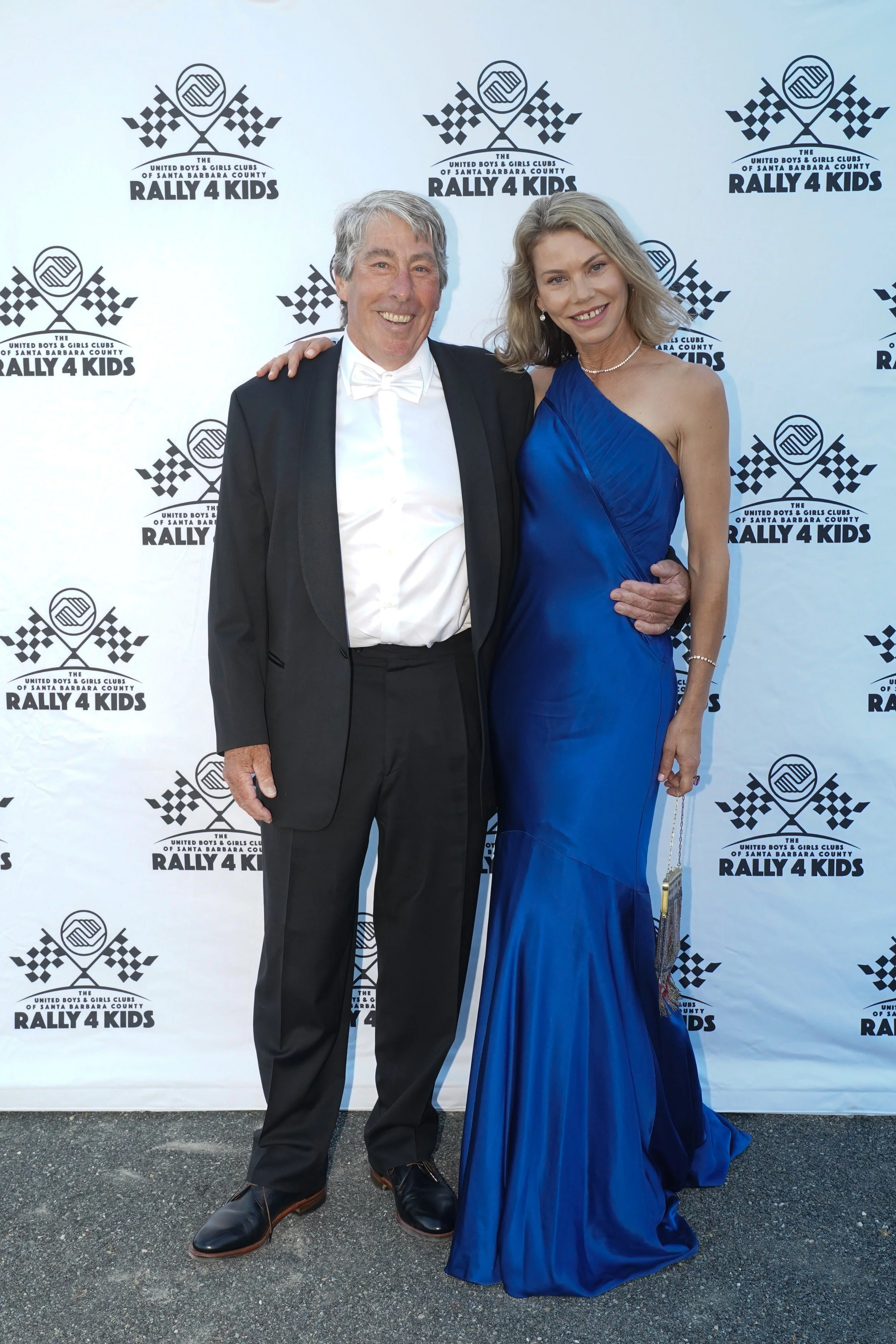 A man in a tuxedo and a woman in a blue evening gown posing together at a charity event called Rally 4 Kids, with a backdrop featuring the event's logo and name.