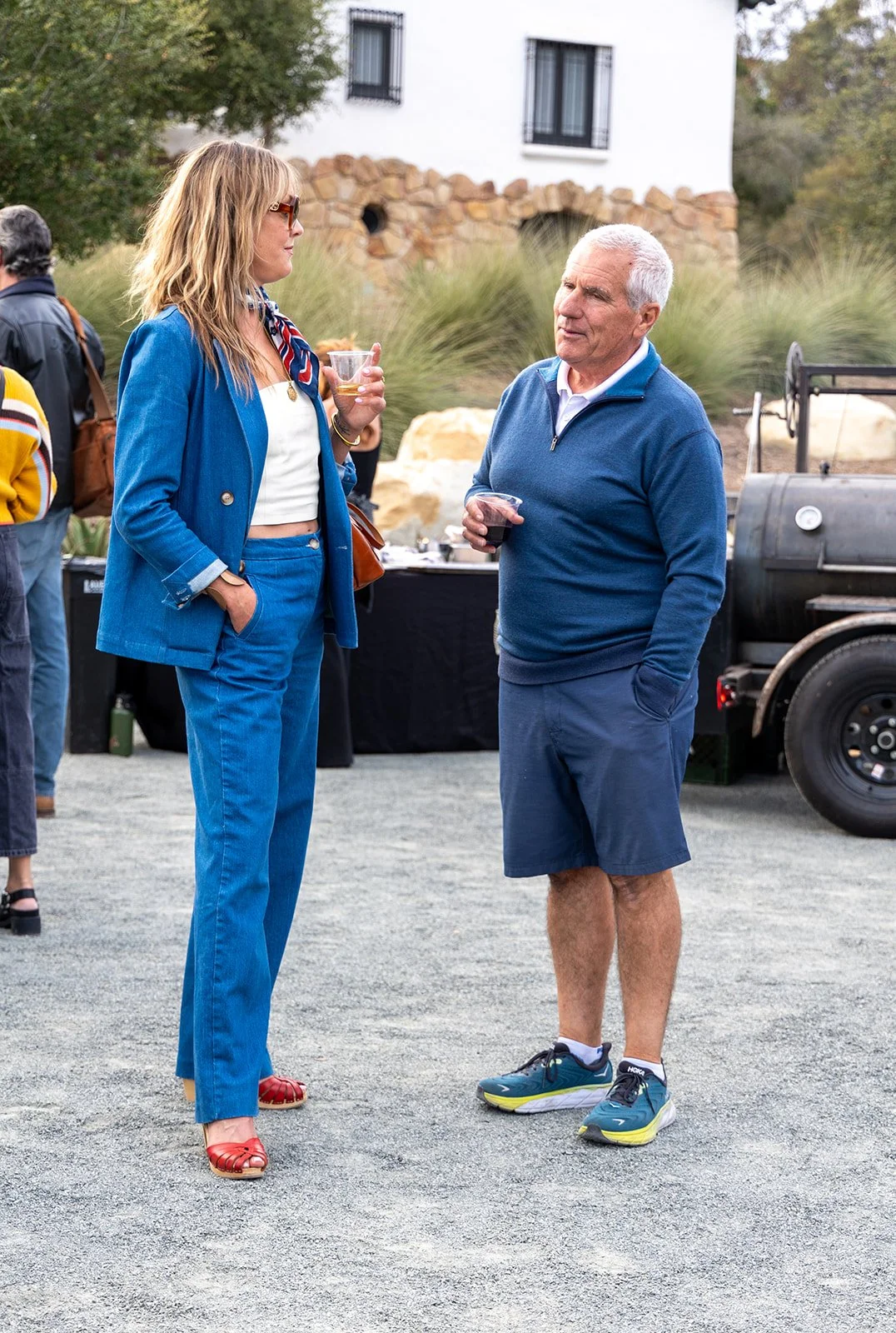 A woman in a bright blue suit with red shoes and sunglasses and a man in a blue pullover and shorts are standing and talking while holding drinks at an outdoor gathering. There are other people and a barbecue grill in the background, along with a whi
