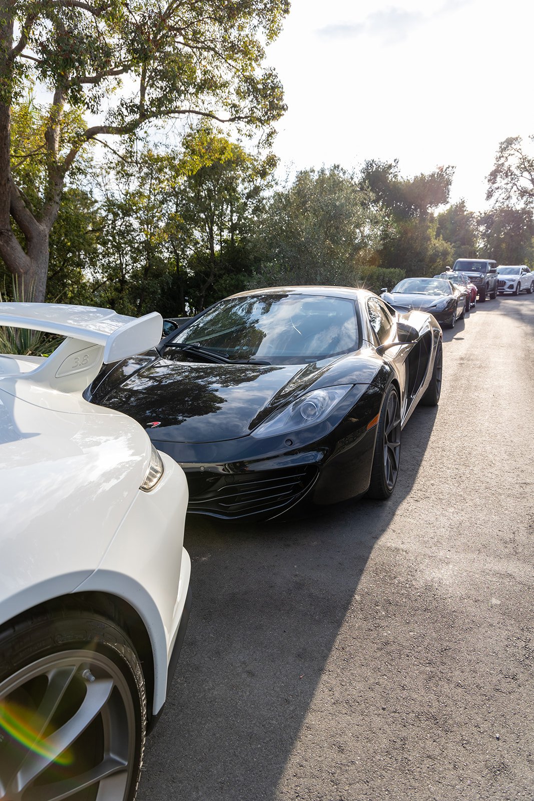 Black sports car parked behind a white sports car on a sunny street with trees and other cars in the background.