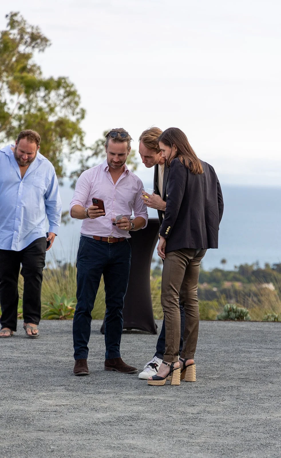 Four people standing outdoors, one man showing something on his phone and another holding a drink, with a scenic background of trees and the ocean.