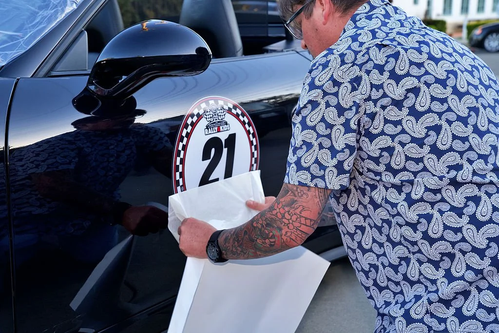 A man with tattoos on his arm and wearing glasses, dressed in a blue and white patterned shirt, affixes a Rally Kids race number 21 sticker onto the door of a black car.
