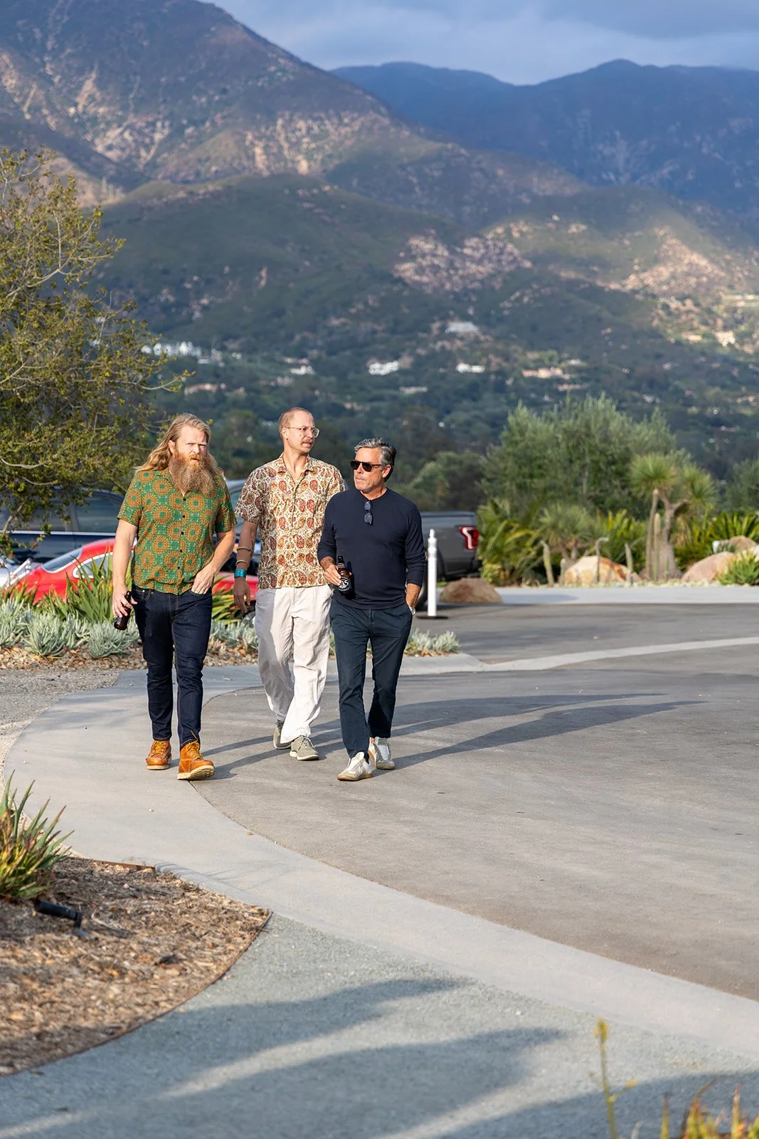 Three men walking outdoors on a curved paved path, with mountains in the background and greenery around, during daytime.