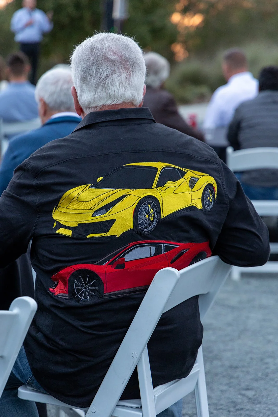 A man sitting on a white chair outdoors, wearing a black jacket with large yellow and red sports car designs on the back, attending an event with other people visible in the background.