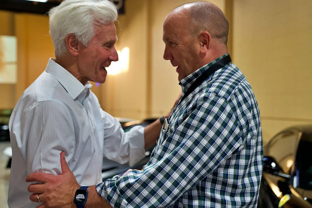 Two middle-aged men smiling and embracing in a warm indoor setting, with one wearing a white shirt and the other in a blue checkered shirt.