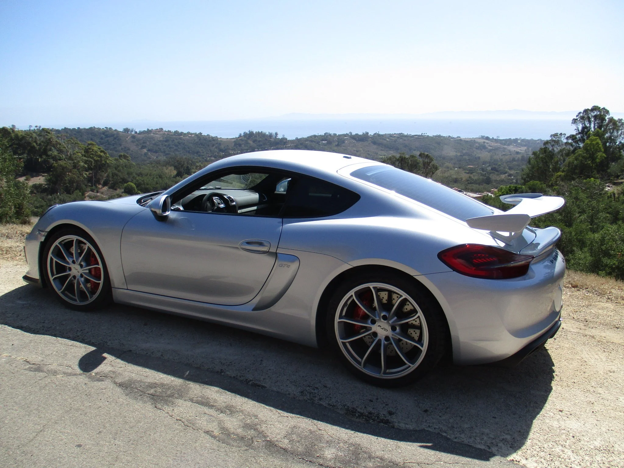 Silver sports car parked on a dirt road with a scenic view of hills, trees, and water in the distance under a clear sky.