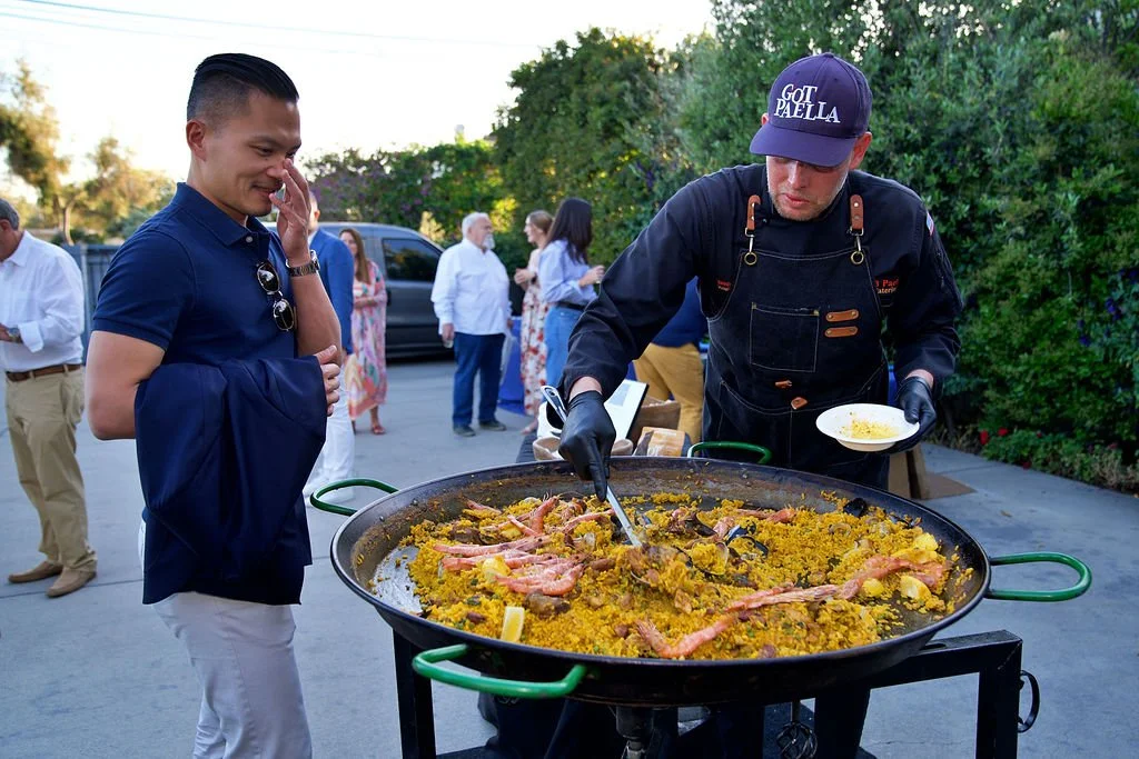 A chef serving seafood paella from a large pan at an outdoor event, with a man in a blue shirt watching and other guests in the background.