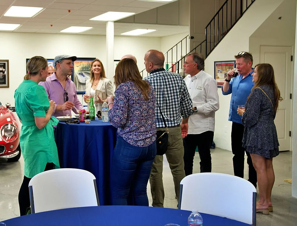 Group of people socializing around a high-top table at an indoor event, with vintage cars visible in the background.