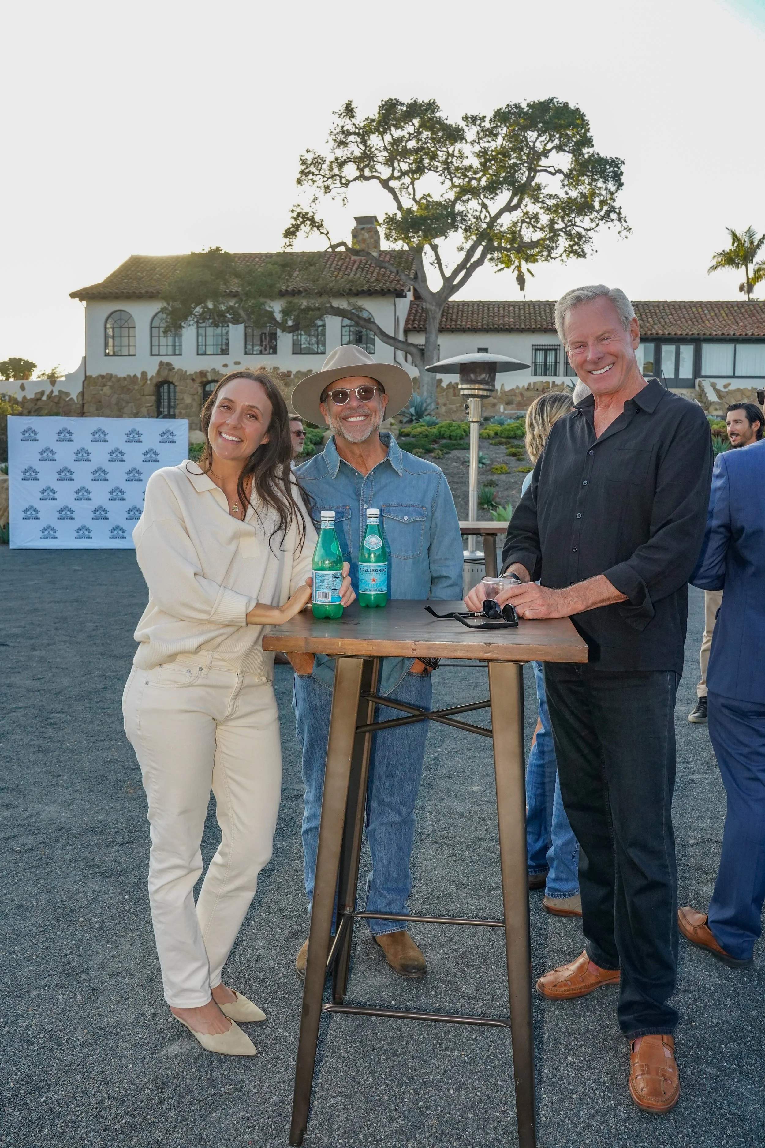 Group of three people standing around a high-top table at an outdoor event, holding bottles of sparkling water, smiling, with a house and trees in the background.
