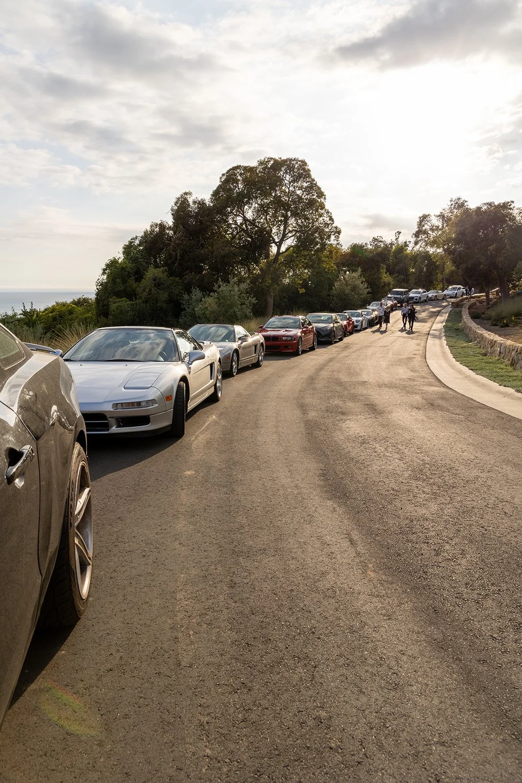 A line of parked cars along a scenic curved road with trees and people walking in the background during the daytime.