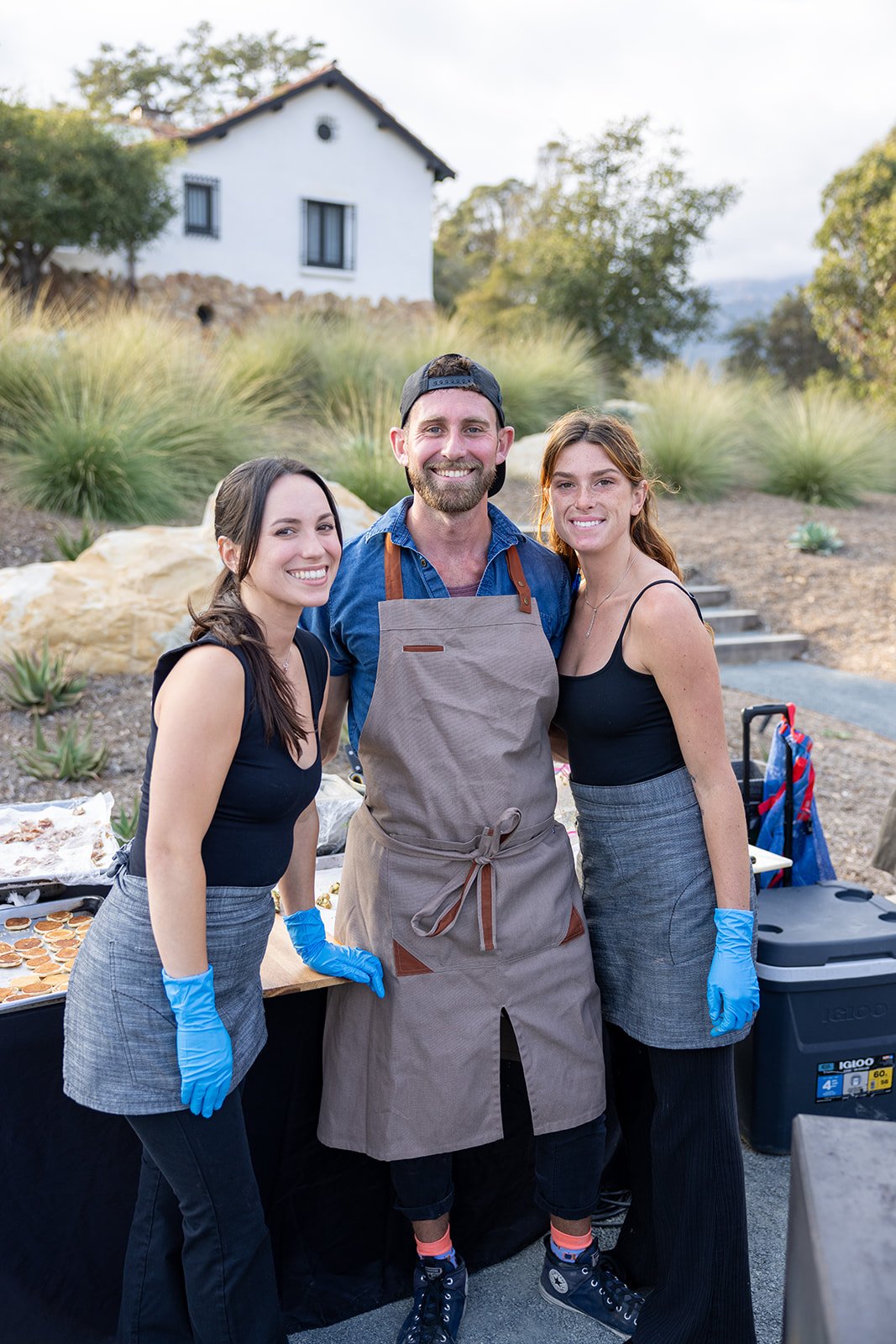 Three smiling people, two women and one man, standing outdoors at a food stall with cookies, wearing black tops, gray skirts, and an apron. They are in a natural setting with plants, rocks, and a house in the background. All are wearing blue gloves.