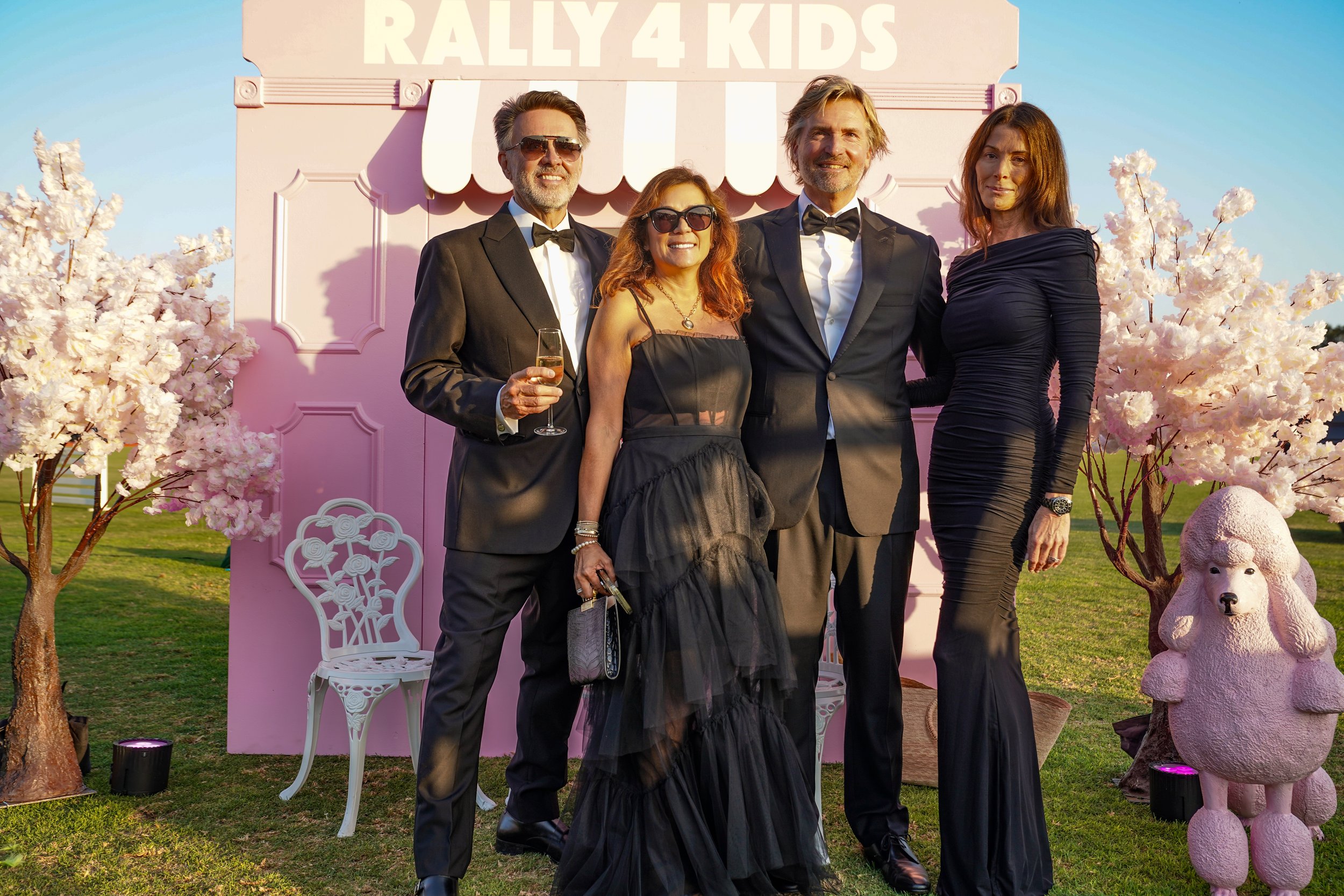 Four adults dressed in formal attire standing outdoors at sunset in front of a pink backdrop that reads "Rally 4 Kids". Two men in tuxedos, two women in black dresses, with pink cherry blossom trees and decorative pink poodle statue on either side.