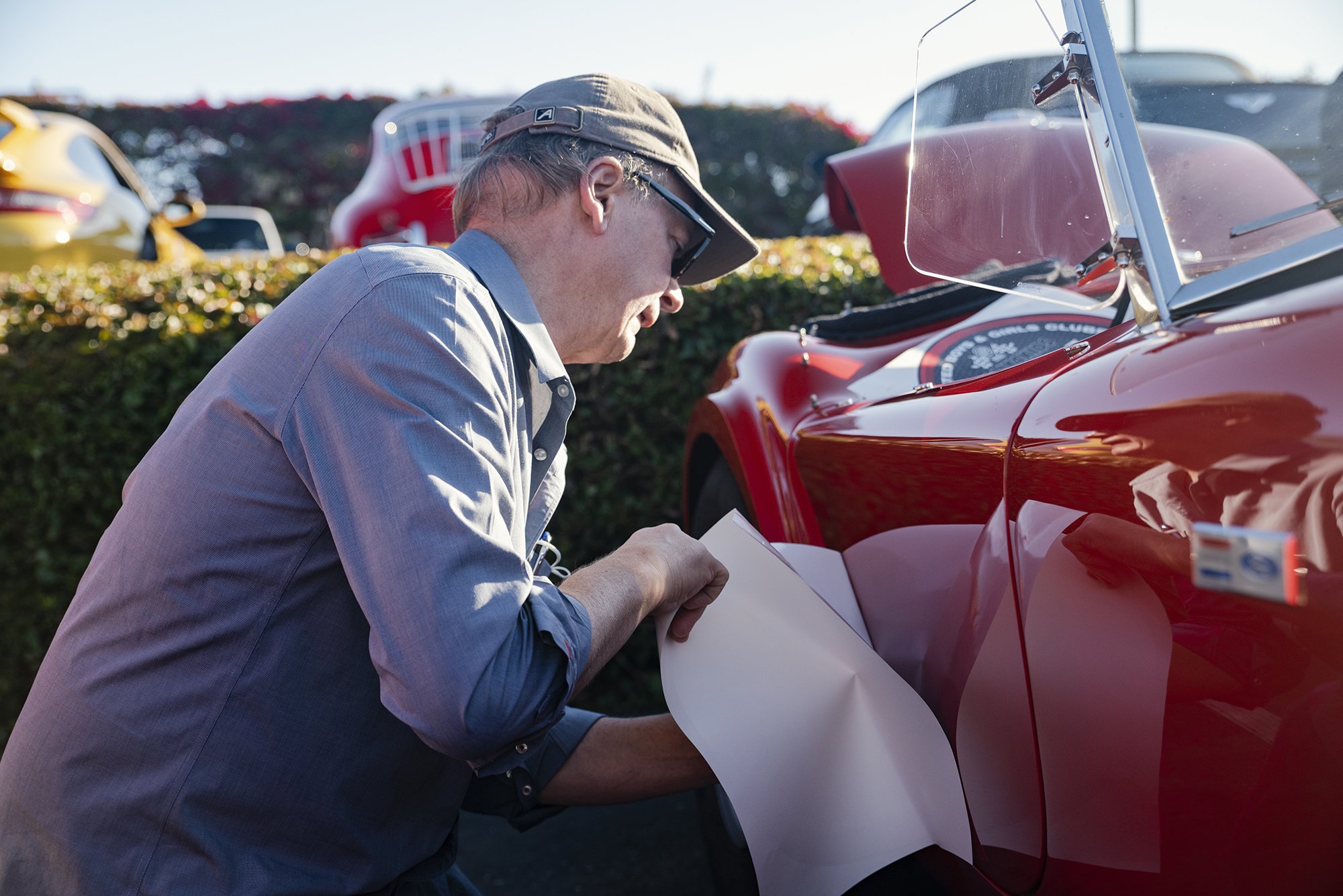 A man wearing a baseball cap and sunglasses applying a sticker to a red vintage race car at a car show.