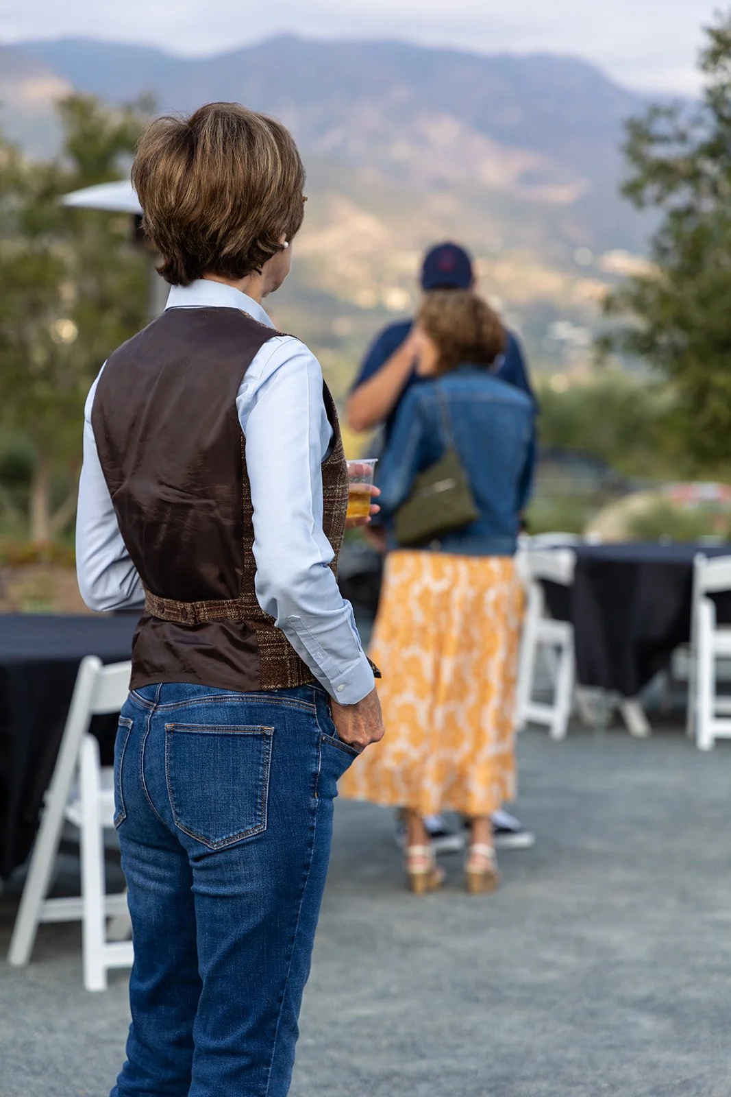 A woman in jeans and a vest standing outside, holding a drink, with two people in the background talking, outdoors with mountains and trees.