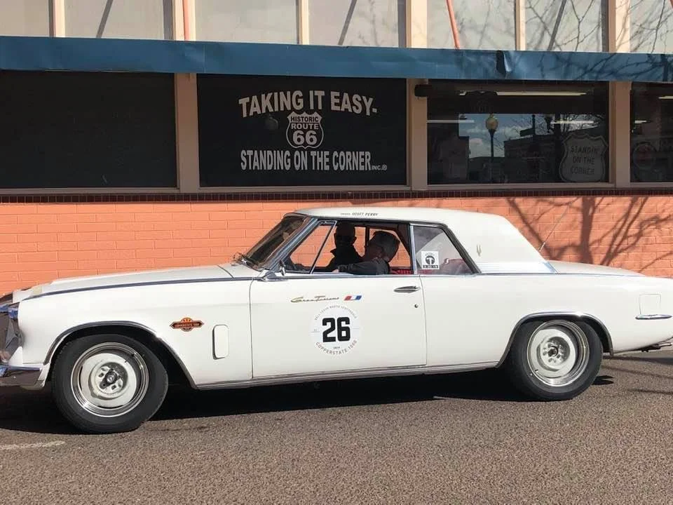A vintage white race car with the number 26 on the door, parked on the street in front of a brick building. Two people are inside, and the background features a sign promoting Route 66.