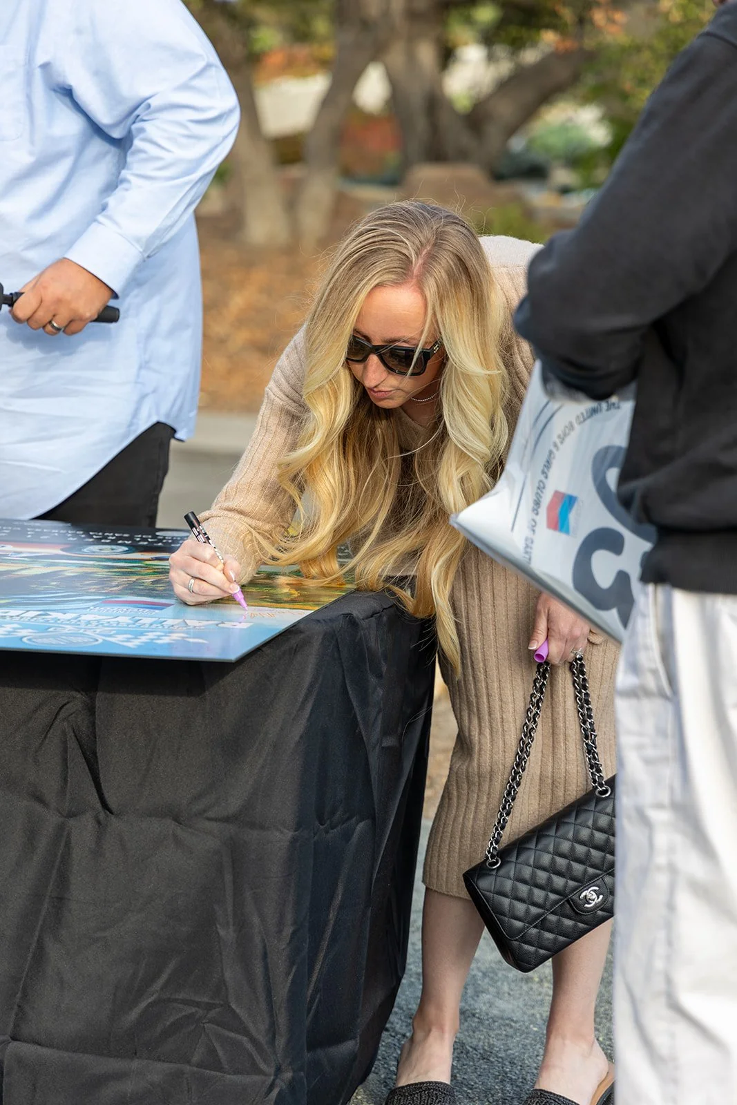 A woman with long blonde hair wearing sunglasses and a beige sweater is signing a poster on a table covered with a black cloth, outdoors during fall. She holds a black purse with a chain strap.
