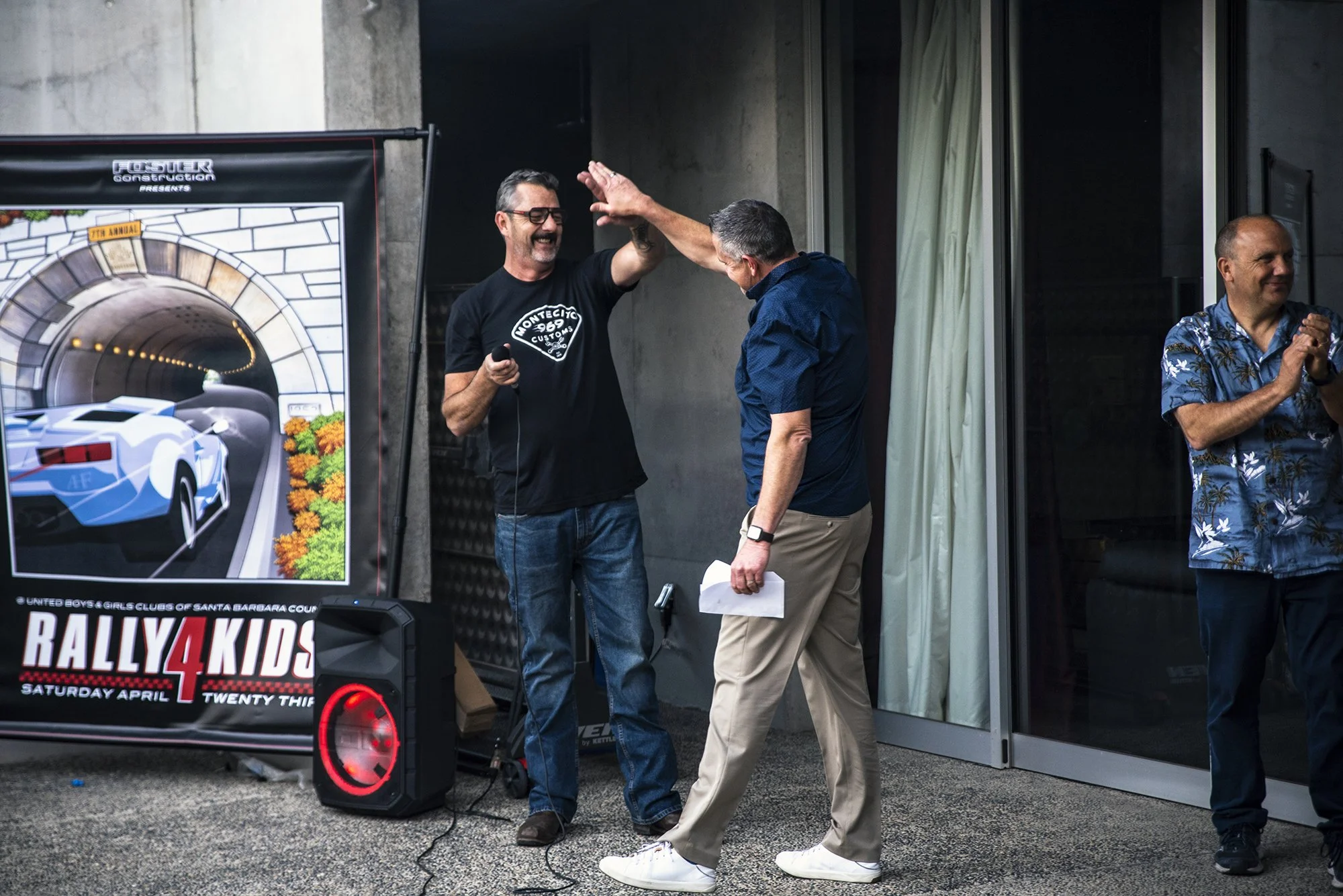 Three men in a room celebrating at an indoor event with a rally car poster in the background. The man on the left is smiling and giving a high five to the man in the middle, who is holding a paper and a microphone. The third man is clapping and stand