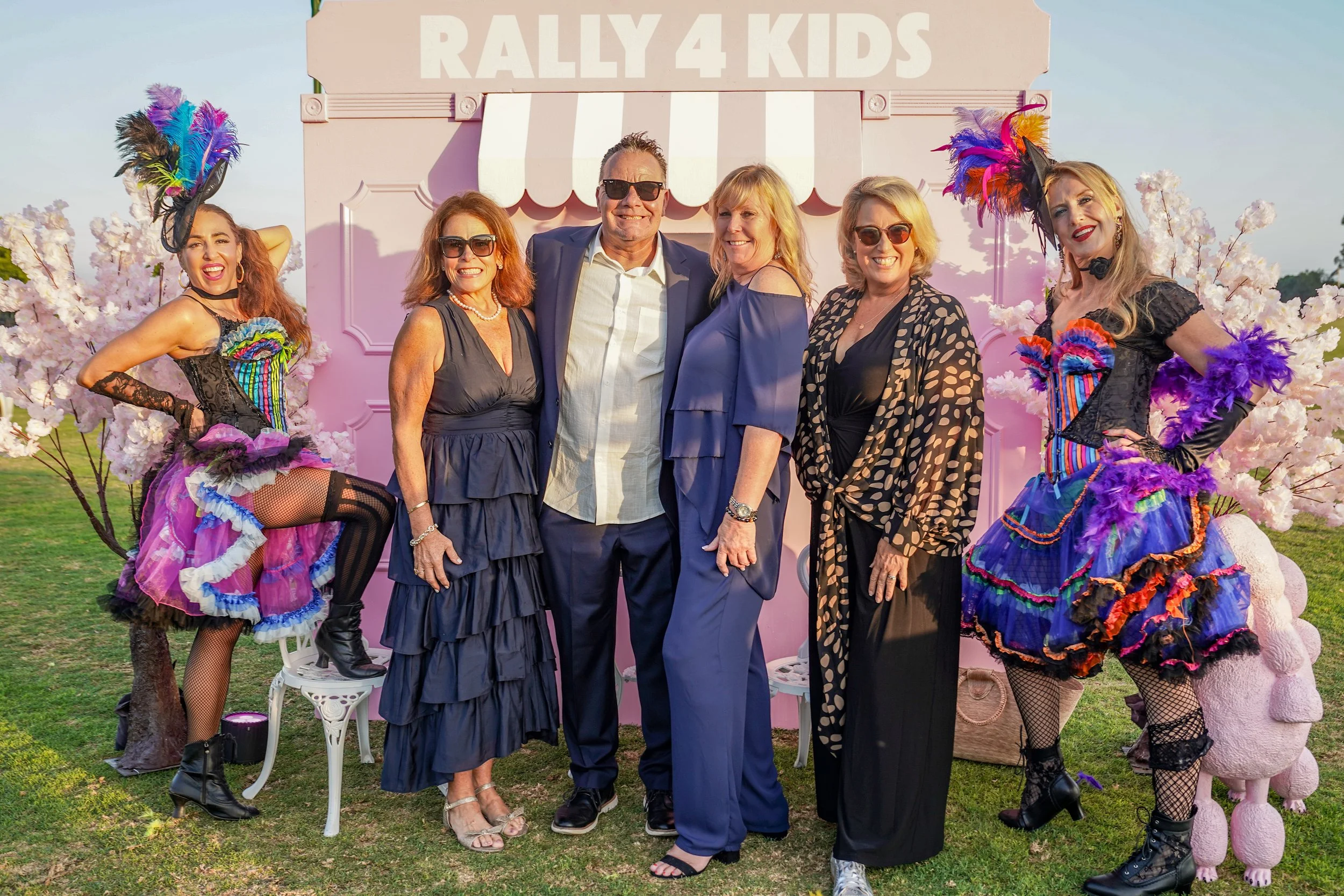 Group of six people dressed nicely standing outdoors in front of a pink backdrop with a sign that says 'RALLY 4 KIDS'. Two women on each side are dressed in colorful, carnival-style costumes with feathered headpieces, while the four people in the mid