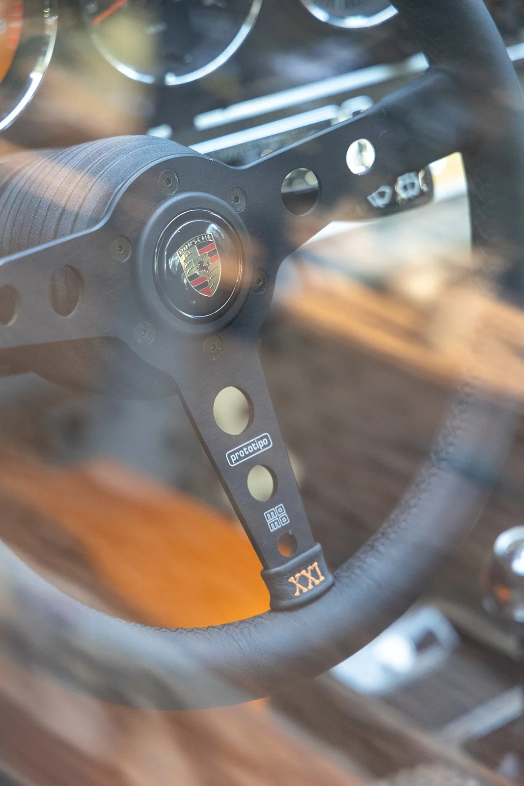 Close-up of a Porsche steering wheel with the Porsche logo at the center, featuring a black metallic finish with drilled holes and branding labels.