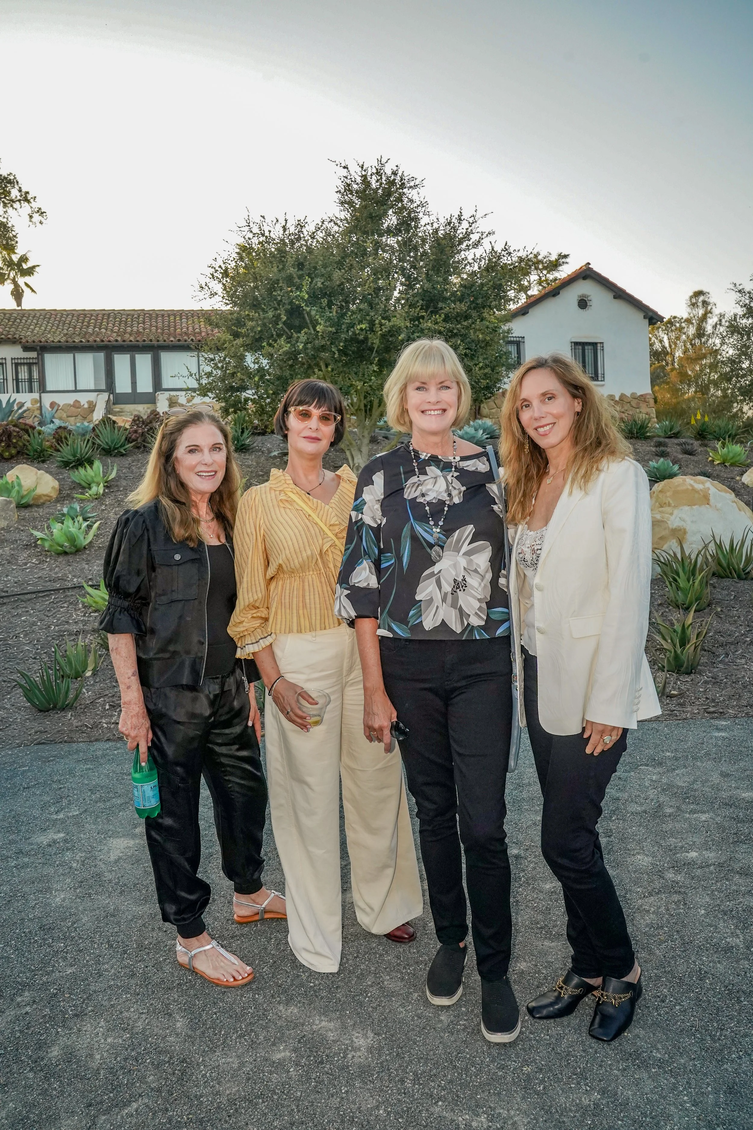Four women posing outdoors in front of a landscaped garden with succulents and a house in the background, during daytime.