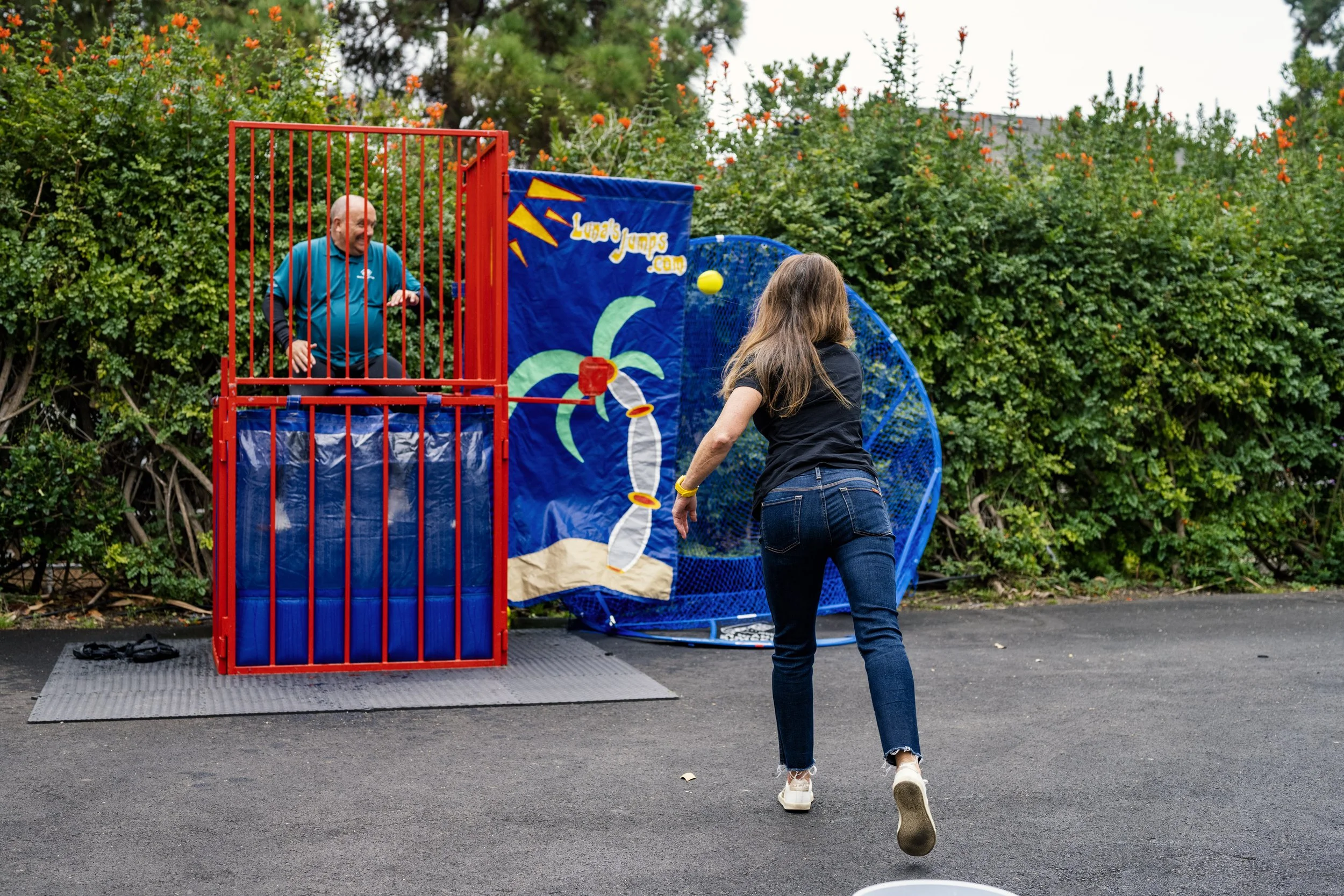 A woman playing baseball with a girl inside a batting cage outdoors, with a backdrop of bushes and trees.
