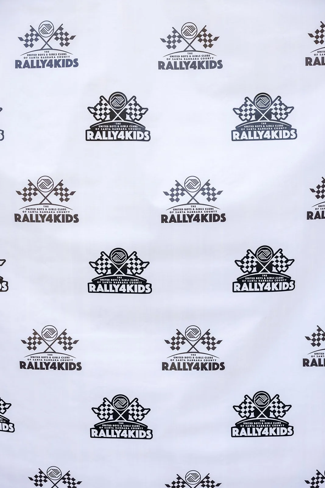 Patterned backdrop with repeating logo for Rally 4 Kids, featuring crossed checkered flags and circular emblem, organized by the United Boys & Girls Clubs of Santa Barbara County.