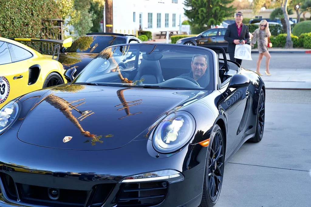 A black Porsche convertible parked on the street with two people inside, one man and one woman in the background walking. There is a yellow race car parked nearby. The street has trees and buildings.