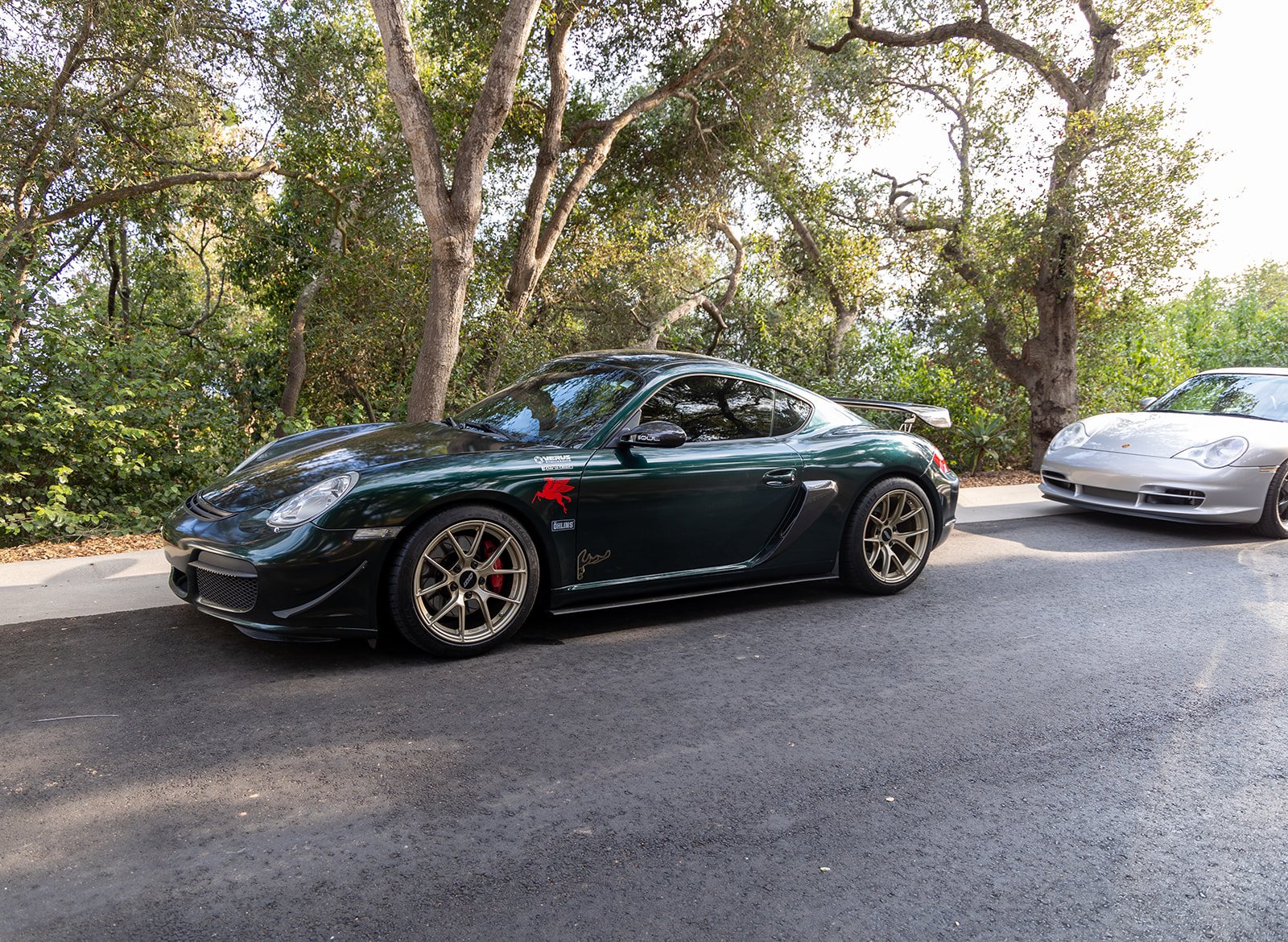 A dark green Porsche 911 sports car parked on the side of a road with another silver sports car behind it, surrounded by trees with green foliage.