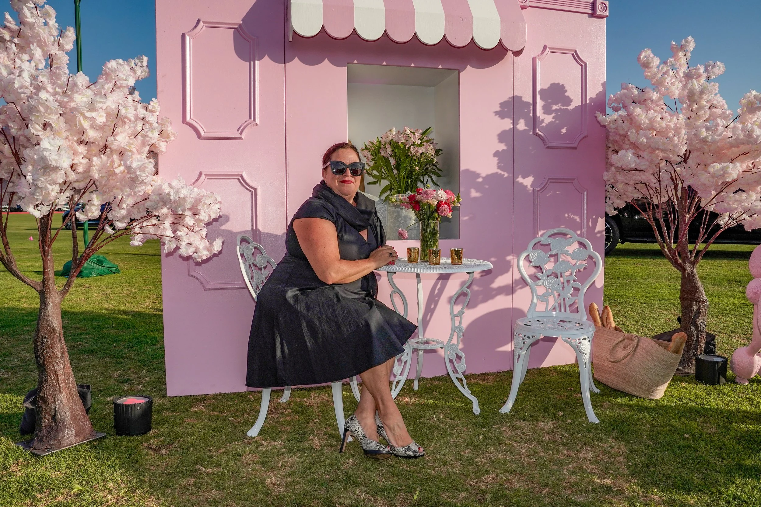 A woman in a black dress and high heels sitting at a white garden table with pink and white flowers in a clear vase, in front of a pink themed backdrop with artificial cherry blossom trees and white chairs, outdoors on a grassy area.