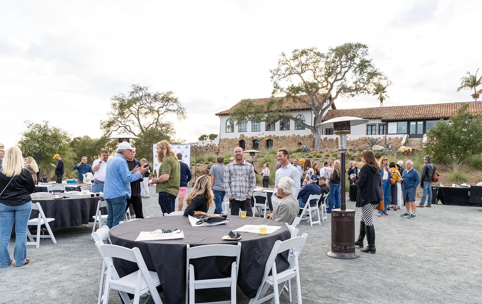 People gathered at an outdoor social event with tables, chairs, and heaters on a gravel surface, with a large house and trees in the background.