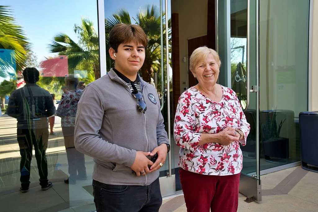 A young man and an older woman standing outside a building with glass doors, reflected in the glass are palm trees and people, suggesting a sunny day.