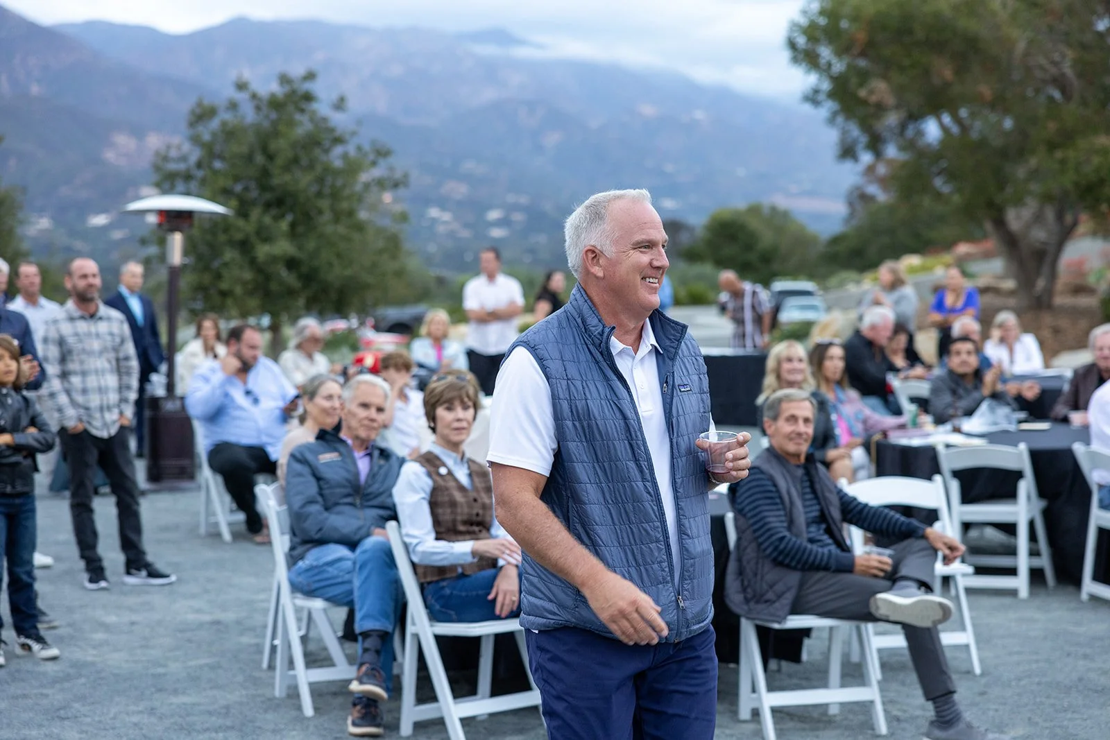 Man in a blue vest holding a plastic cup smiling at outdoor gathering with seated and standing guests in a scenic mountainous area.